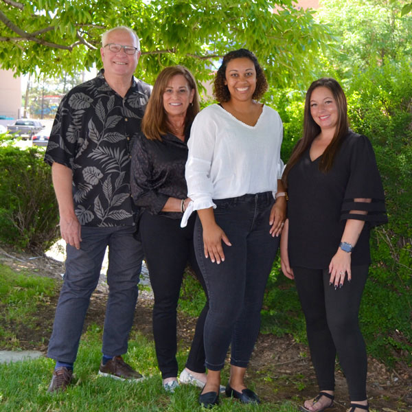 The image shows four individuals posing together outdoors during daylight hours, with a person standing at the center front who appears to be smiling and wearing a white top, flanked by two other individuals on either side of varying heights and genders, and an older adult male standing behind them all.