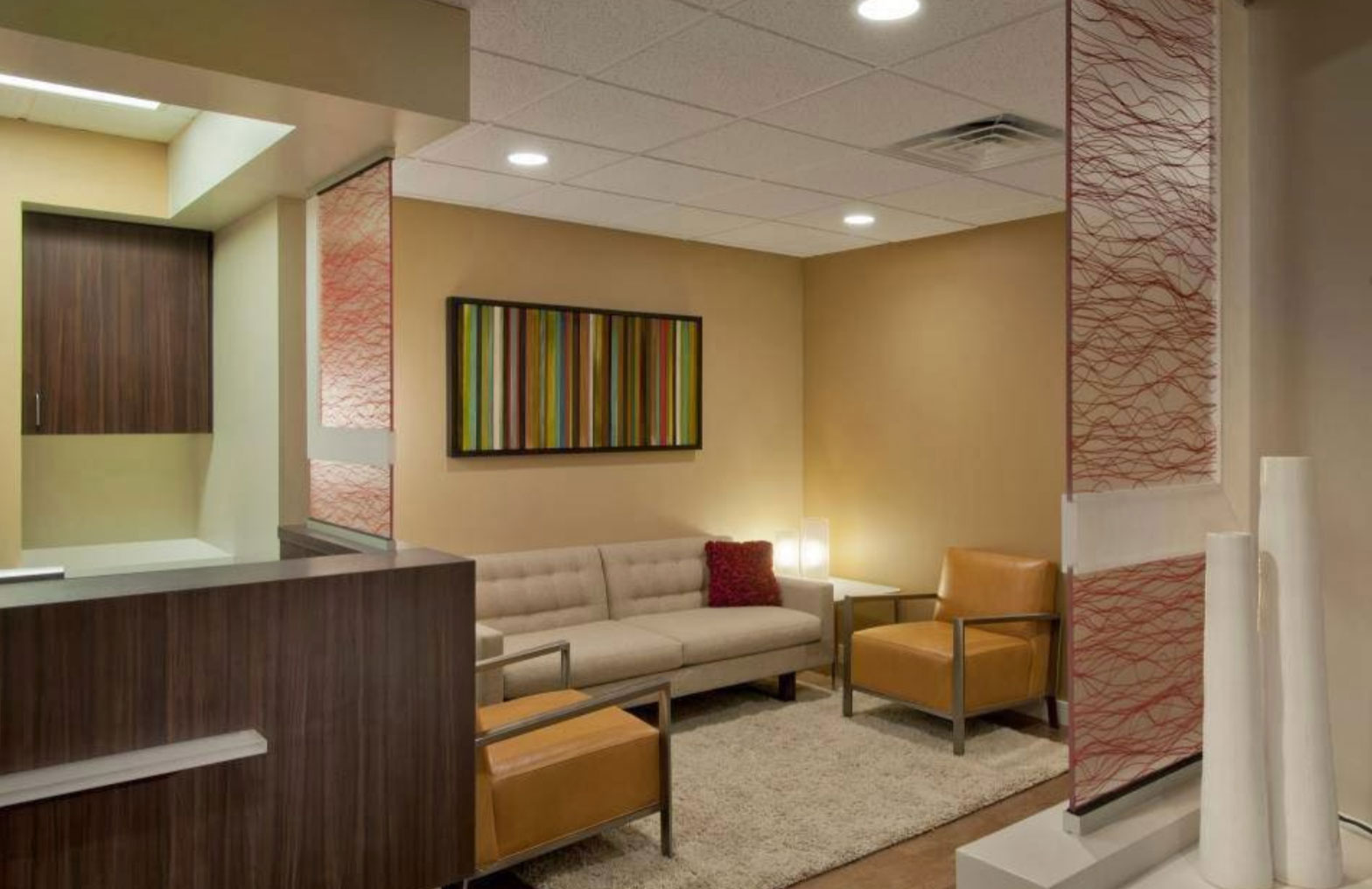 The image shows a modern interior space with a contemporary design featuring a large glass wall separating two areas, one side with a seating area and a desk, and the other with a couch and a coffee table, all set against a backdrop of neutral tones and colorful accent pieces.