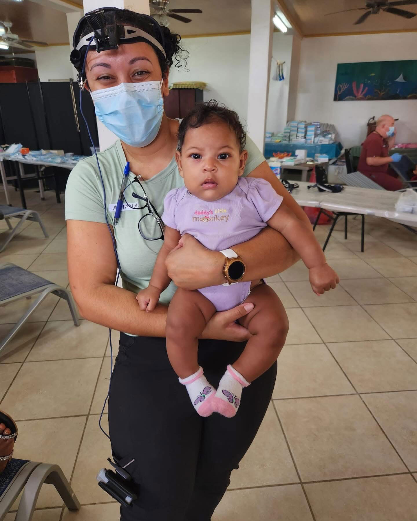 In the image, there is a woman holding a baby, both wearing masks. They are indoors, possibly at a medical facility or an emergency shelter, given the presence of cots and medical equipment in the background. The woman appears to be a healthcare professional, as suggested by her attire and the context.