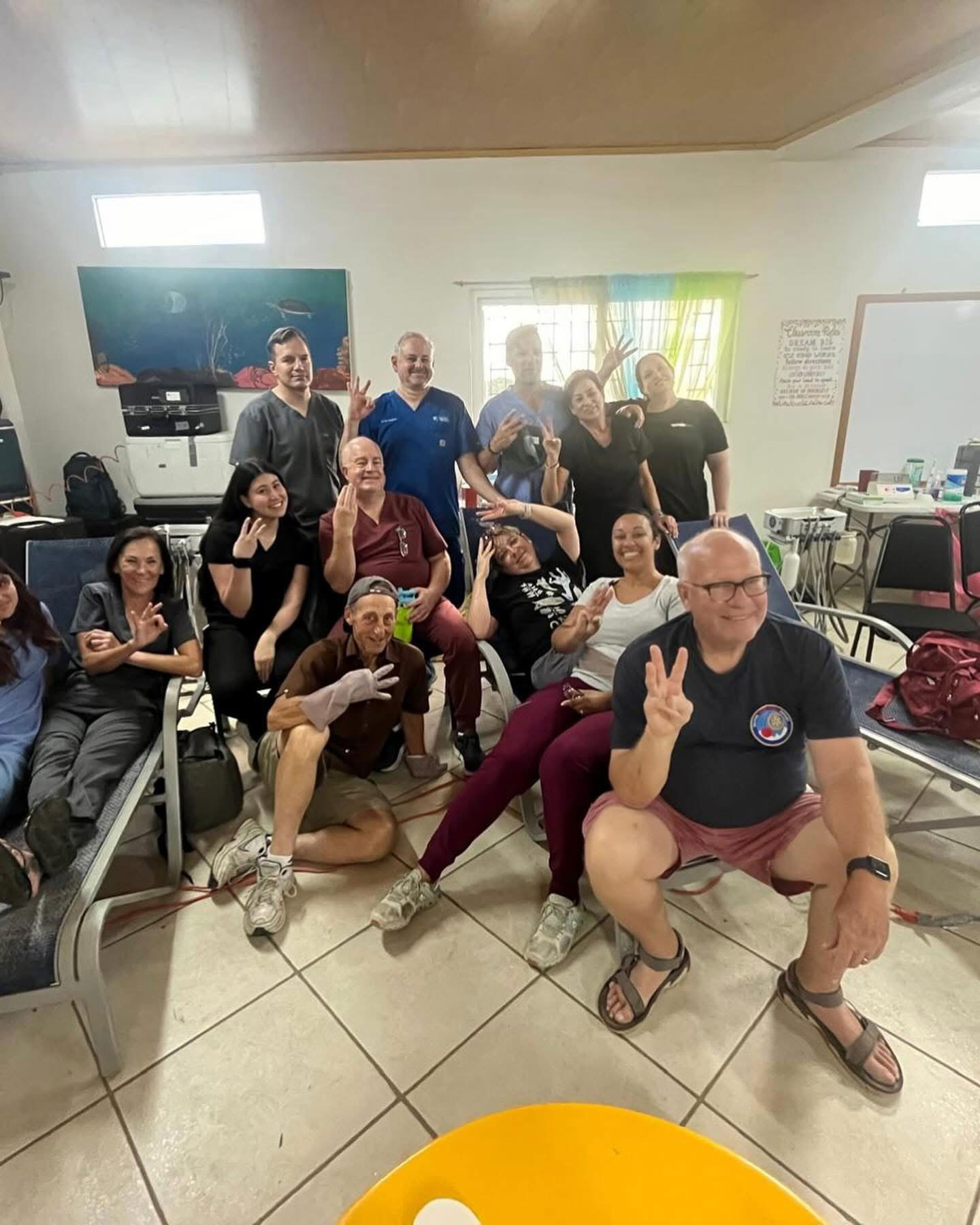A group of people posing together in an indoor setting, some holding up peace signs, with chairs and a table visible.